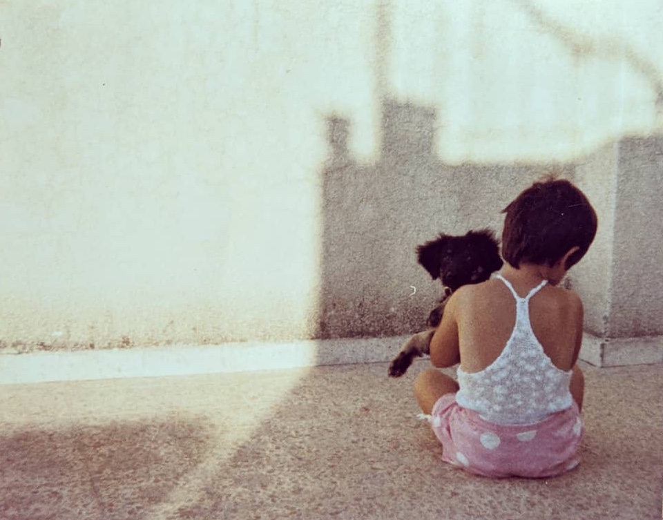 A 4 year-old Esti photographed from the back while sitting on the floor during a summer day, holding a small puppy named Chompi